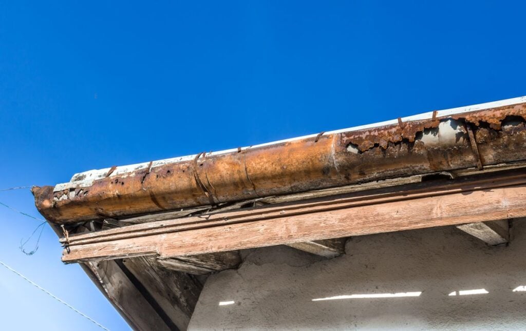 A rusty, damaged rain gutter is attached to the edge of a roof on a building, with a clear blue sky in the background.