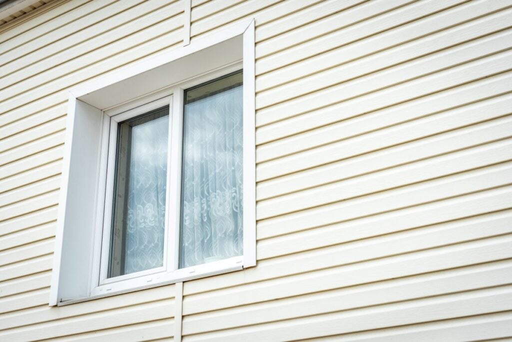 A white-framed window on the side of a house with cream-colored horizontal vinyl siding. The window has sheer curtains inside and the exterior is clean and well-maintained.