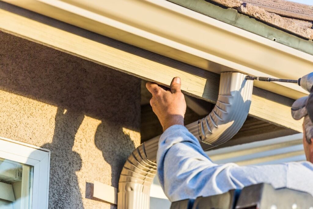 A person installs a white rain gutter system on the roof edge of a house, securing it with screws while standing on a ladder in bright sunlight.