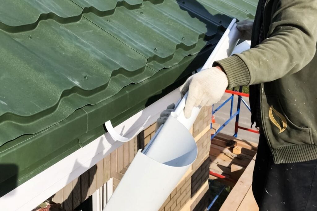 A person wearing gloves installs a white rain gutter on the edge of a green metal roof, standing on scaffolding next to a brick building.