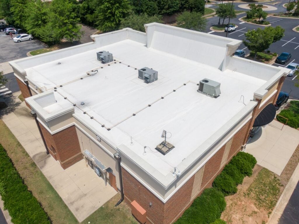Aerial view of a flat white TPO rooftop on a brick commercial building with multiple HVAC units and vents; surrounded by sidewalks, parking lots, trees, and landscaped greenery.