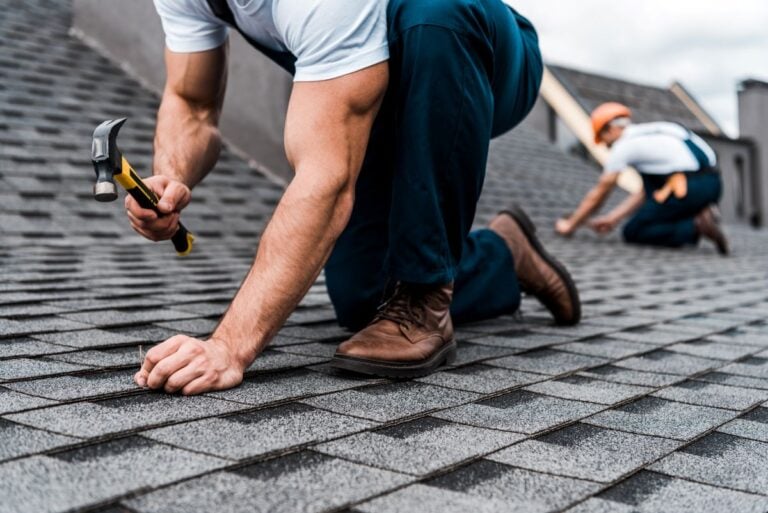 Two workers in uniforms and boots install shingles on a roof. One is close-up, kneeling and using a hammer, while the other is blurred in the background, also working on the roof.