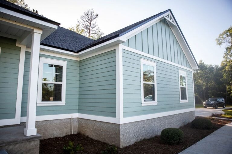 Light blue house with white trim and a dark roof, featuring two windows and landscaping with small bushes. A car is parked on the street in the background on a sunny day.