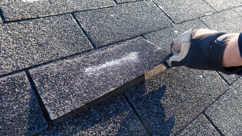 A gloved hand lifts a roof shingle marked with a white chalk line, revealing the shingles underneath.