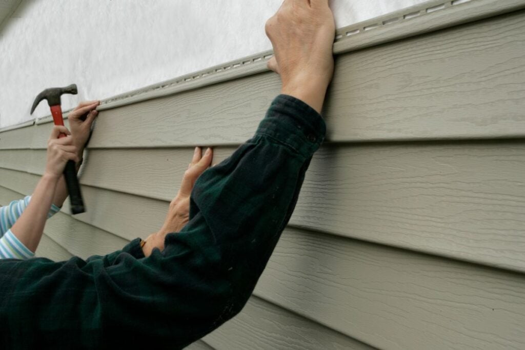 Two people install beige vinyl siding on a house. One person holds the siding in place, while the other uses a hammer to secure it with nails. Only their arms and hands are visible.