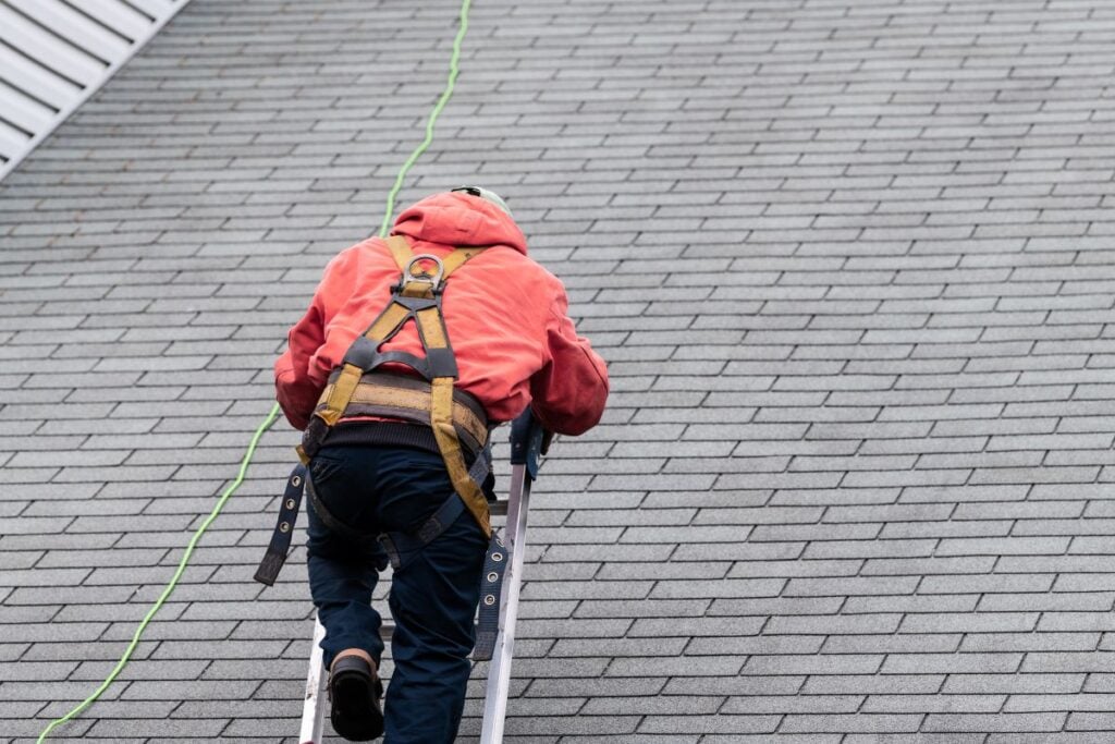 A person wearing a red jacket and safety harness climbs a ladder onto a gray shingled roof, with a safety rope attached above.