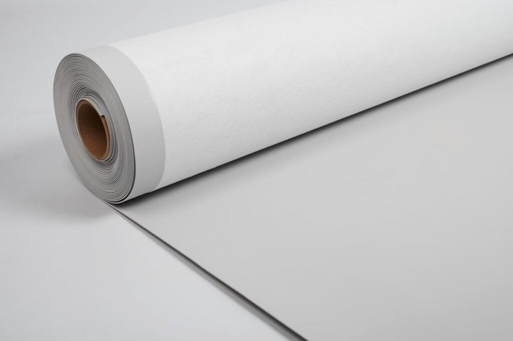 A large roll of white TPO material lies partially unrolled on a smooth, light gray surface, with the roll’s brown cardboard core visible on one end.