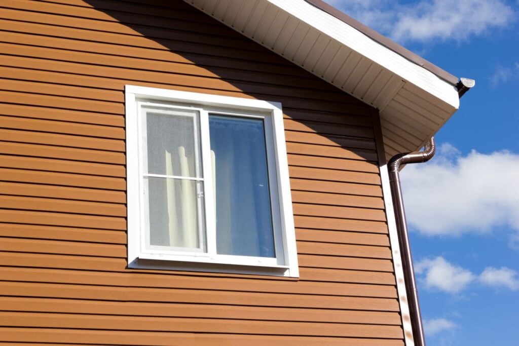 Close-up of a brown house’s upper story with white-trimmed window and visible gutter, under a blue sky with clouds.