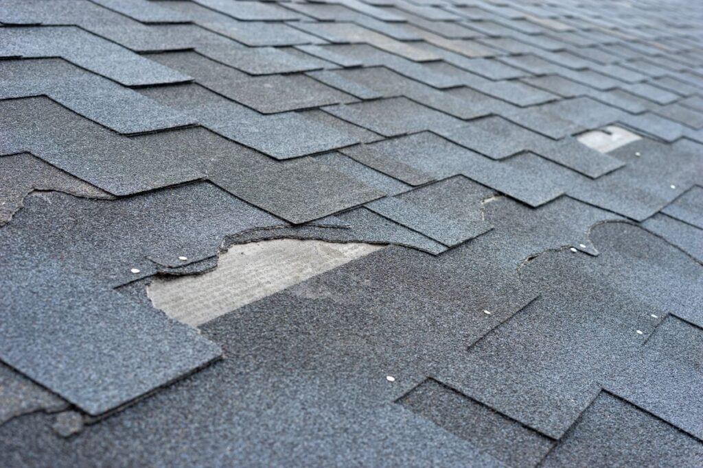 Close-up of a roof with dark asphalt shingles, some of which are damaged or missing, exposing the underlying material.