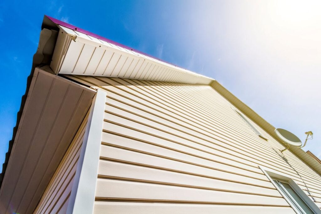 Low-angle view of a house exterior with light-colored siding, a white corner trim, and a satellite dish mounted on the wall.