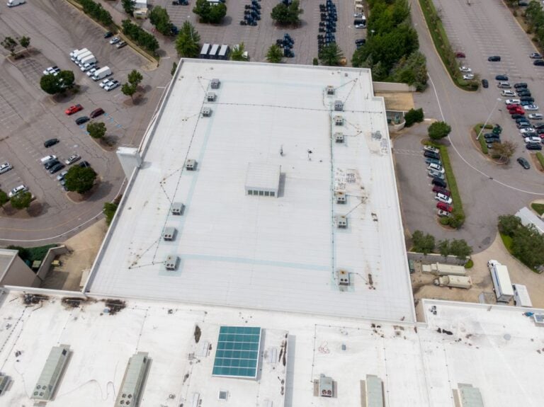 Aerial view of a large flat commercial building roof with multiple HVAC units and a skylight, surrounded by parking lots with scattered vehicles.
