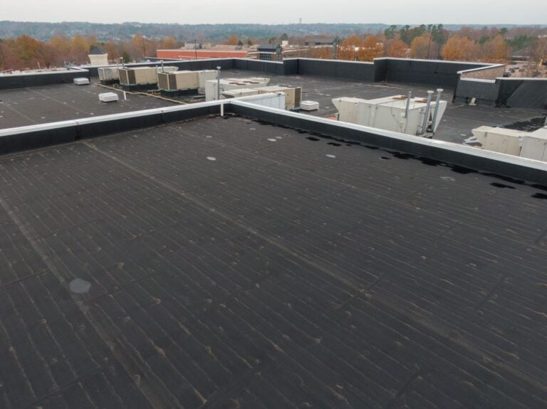 A flat, dark-colored commercial roof with several HVAC units and a view of trees and buildings in the distance.
