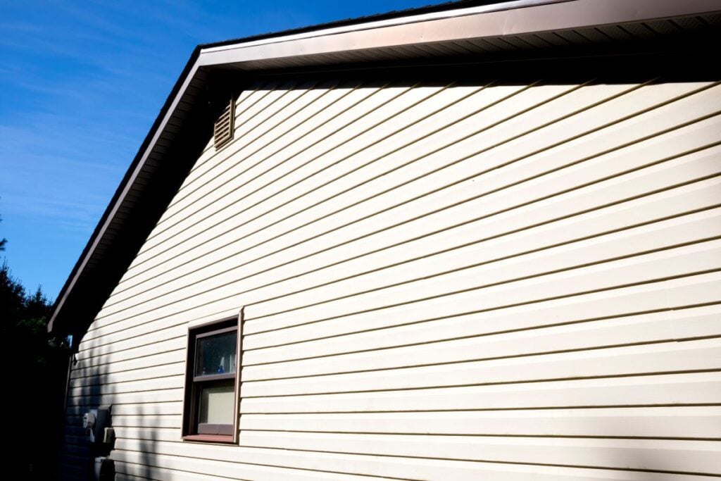 The side of a beige-sided house with a single window under a peaked roofline against a blue sky.