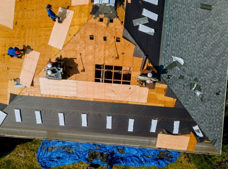 Aerial view of workers installing plywood sheathing and roofing materials on a house roof.