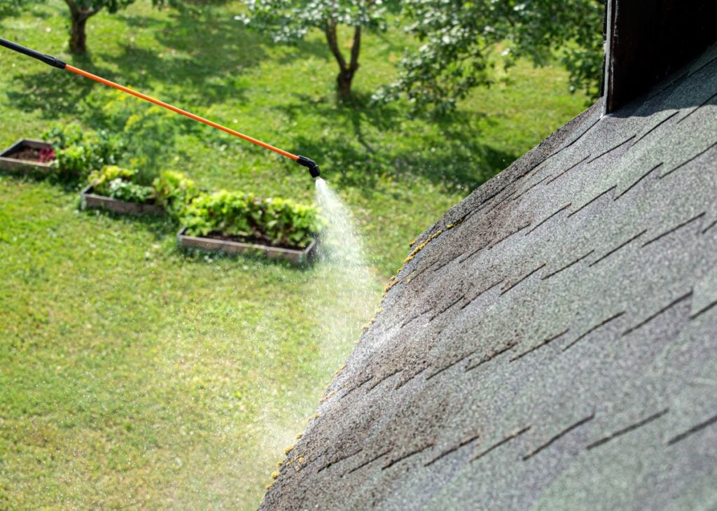A person using a long-reach spray wand to apply cleaning solution to a section of an asphalt shingle roof.