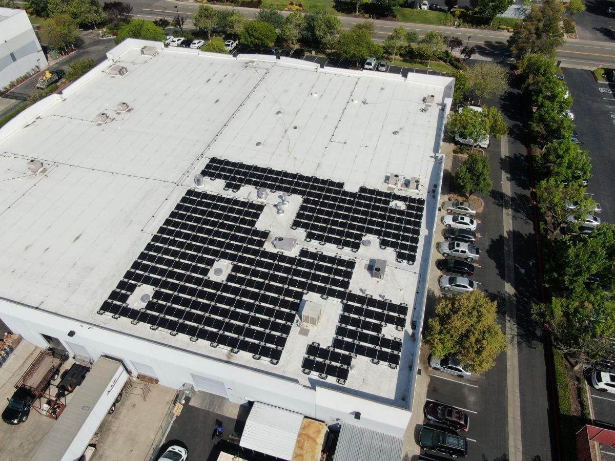 Aerial view of a commercial building with a large grid of solar panels on its flat roof and a parking lot to the side.