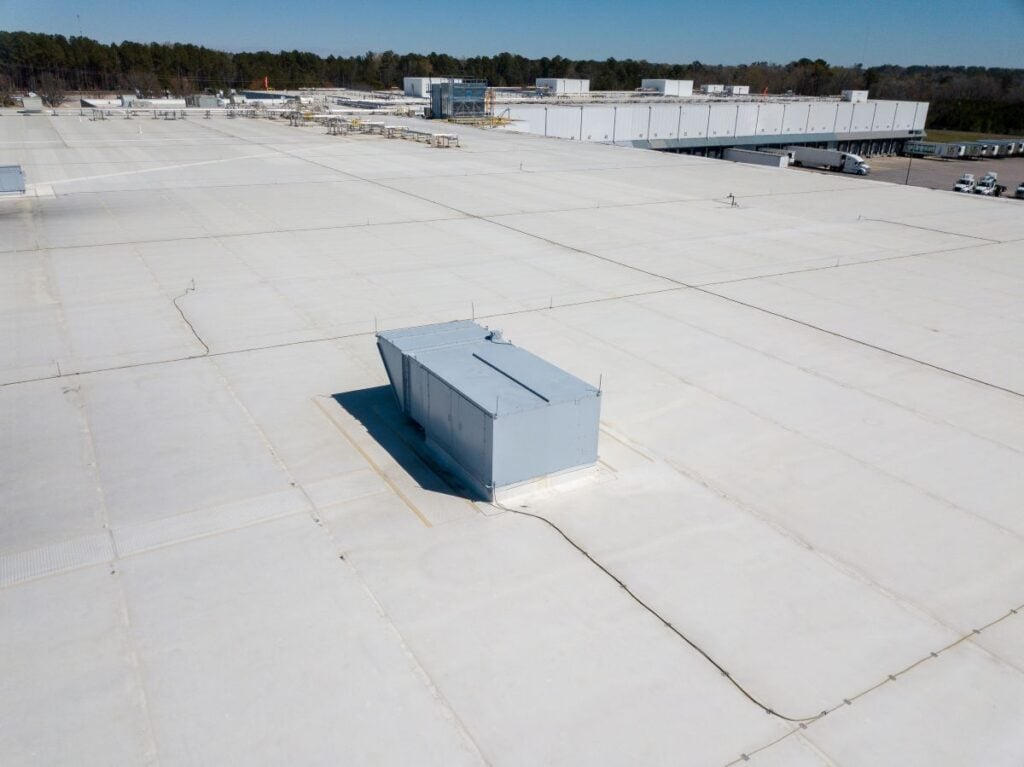 Aerial view of a large white flat industrial warehouse roof with a grey HVAC unit in the center. TPO vs. PVC Roofing