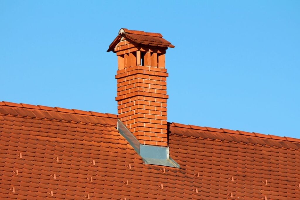 A brick chimney with a small peaked roof cap rising from a tiled roof against a clear blue sky.