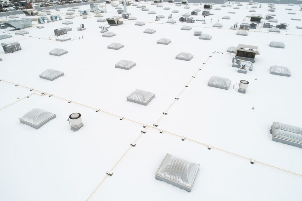 Aerial view of a flat building roof covered in snow, punctuated by numerous square skylights and HVAC units. Commercial Roofing Systems.