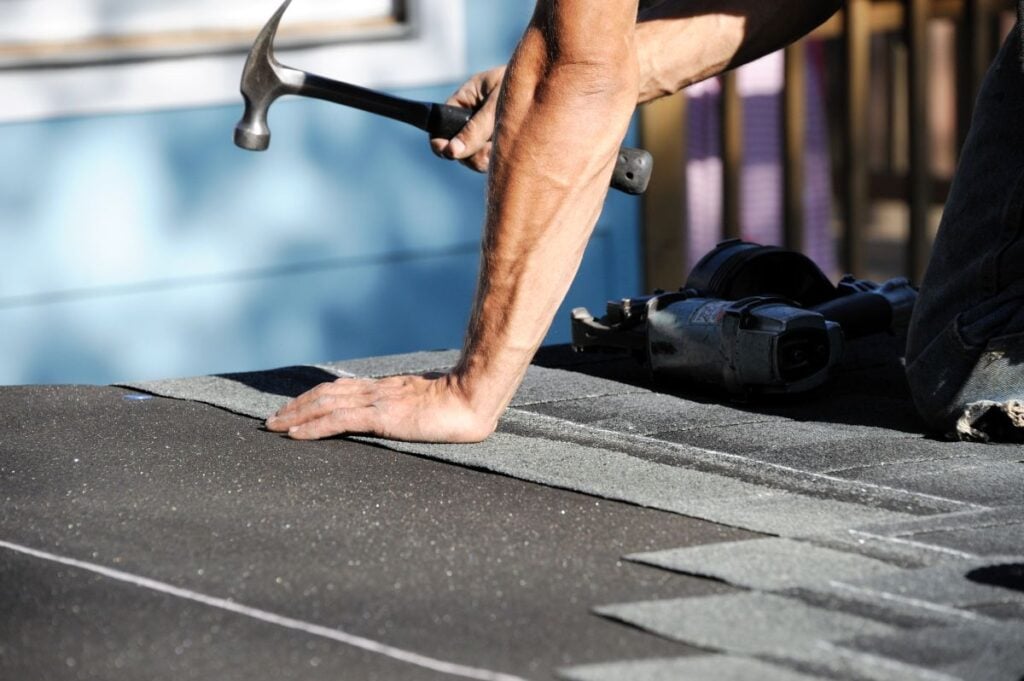 A roofer holding a hammer while installing asphalt shingles on a roof.