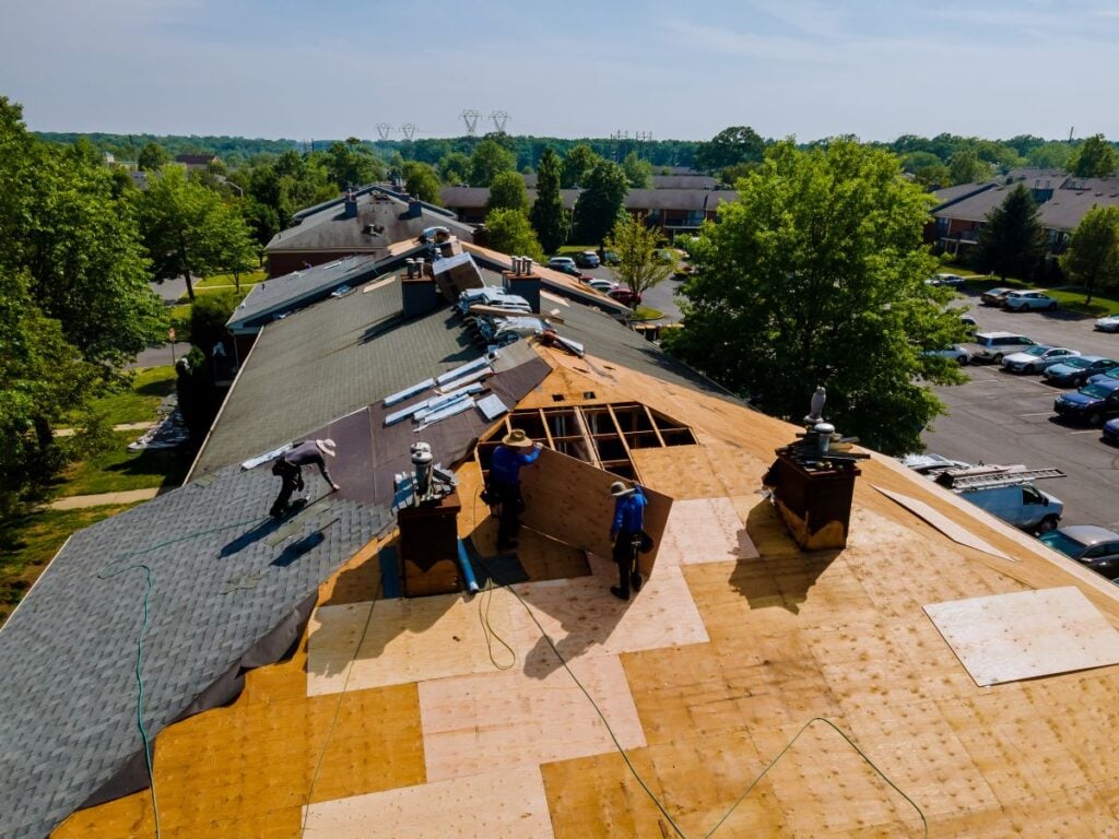 Three construction workers repair a residential roof, installing new plywood sheeting over the roof frame.