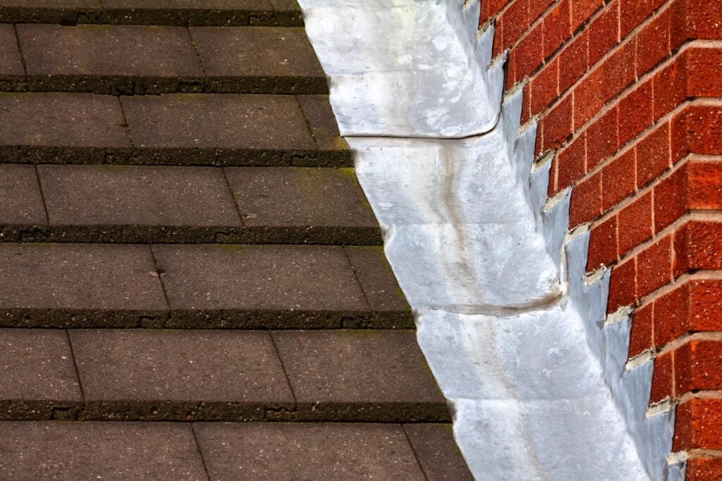 A close-up of dark roof tiles meeting a red brick wall, separated by a curved metal flashing strip.